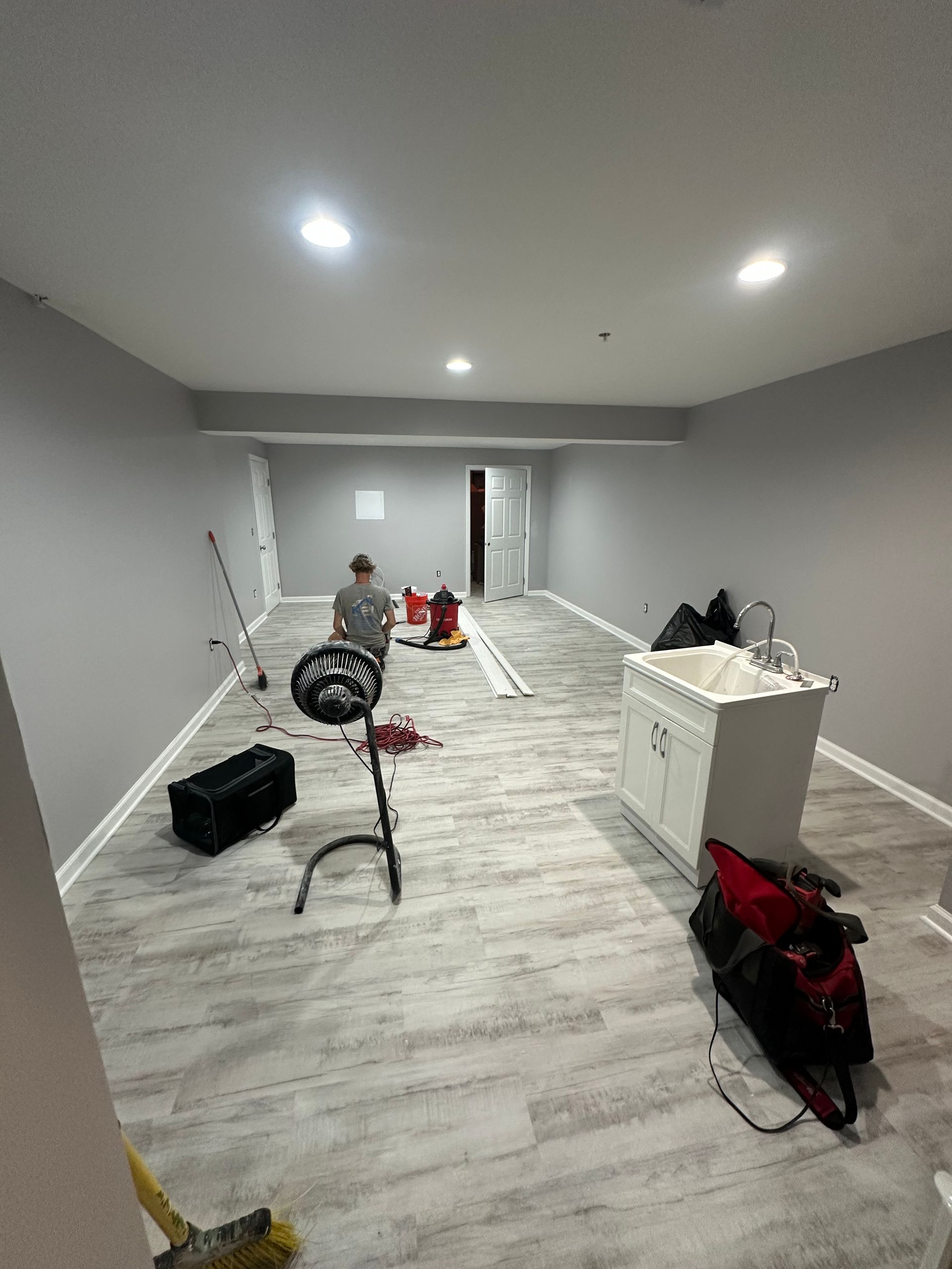 Basement renovation: light gray walls, wood-look flooring, sink cabinet, tools scattered, person in the background.