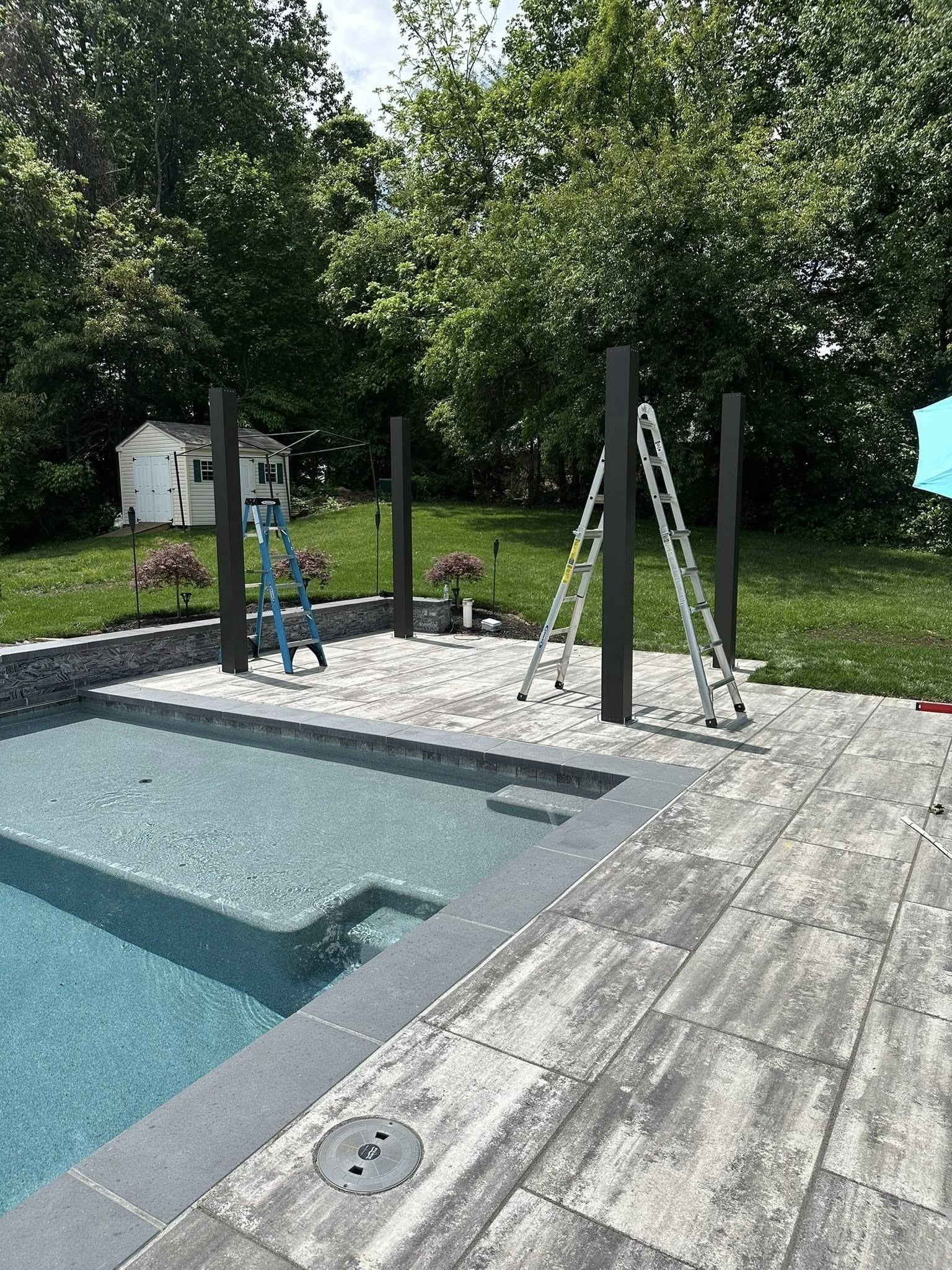 Poolside construction with black posts and ladders, trees, and blue pool water.