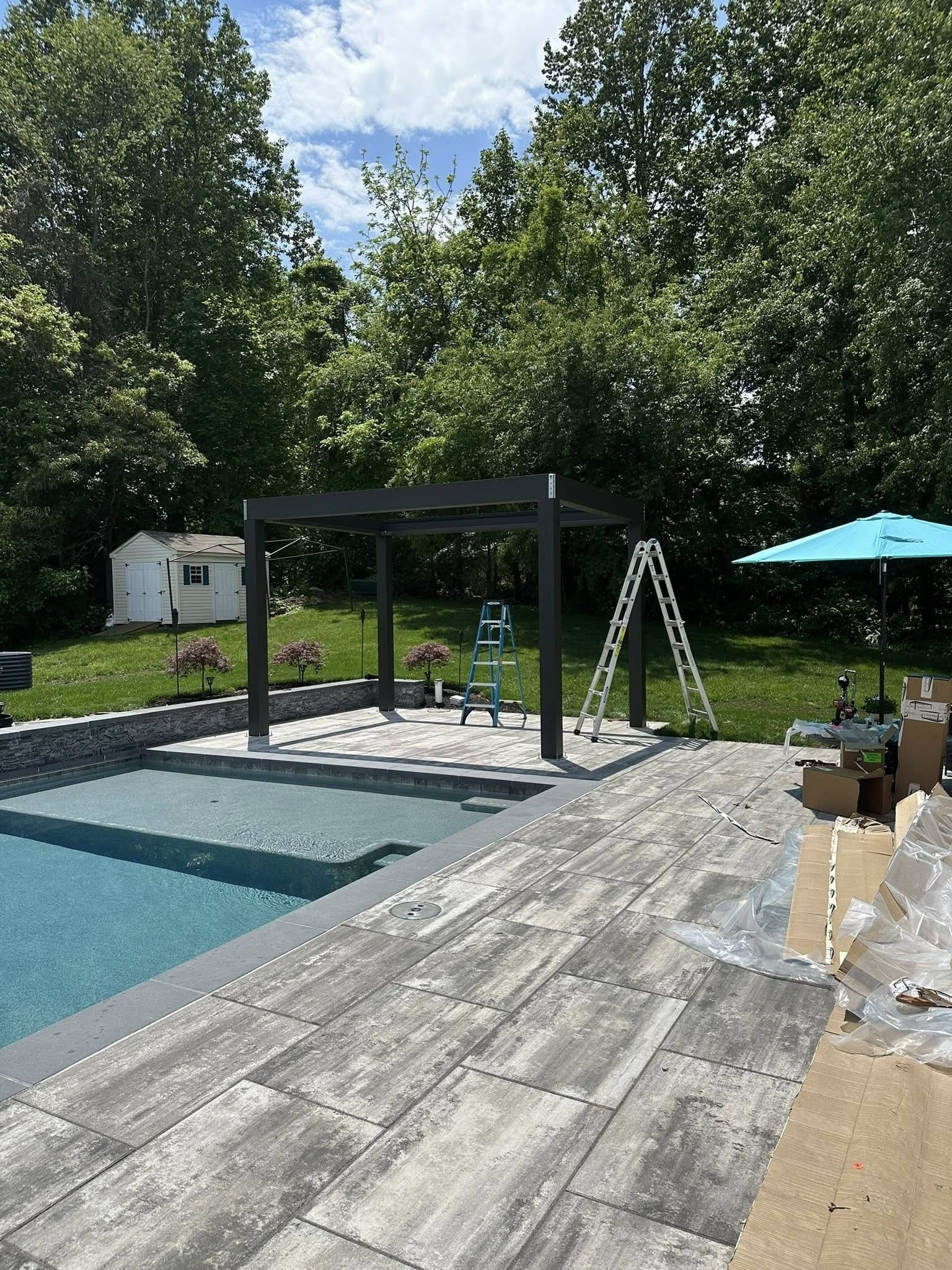 Poolside patio with dark pergola, ladder, blue umbrella, and shed, surrounded by trees.