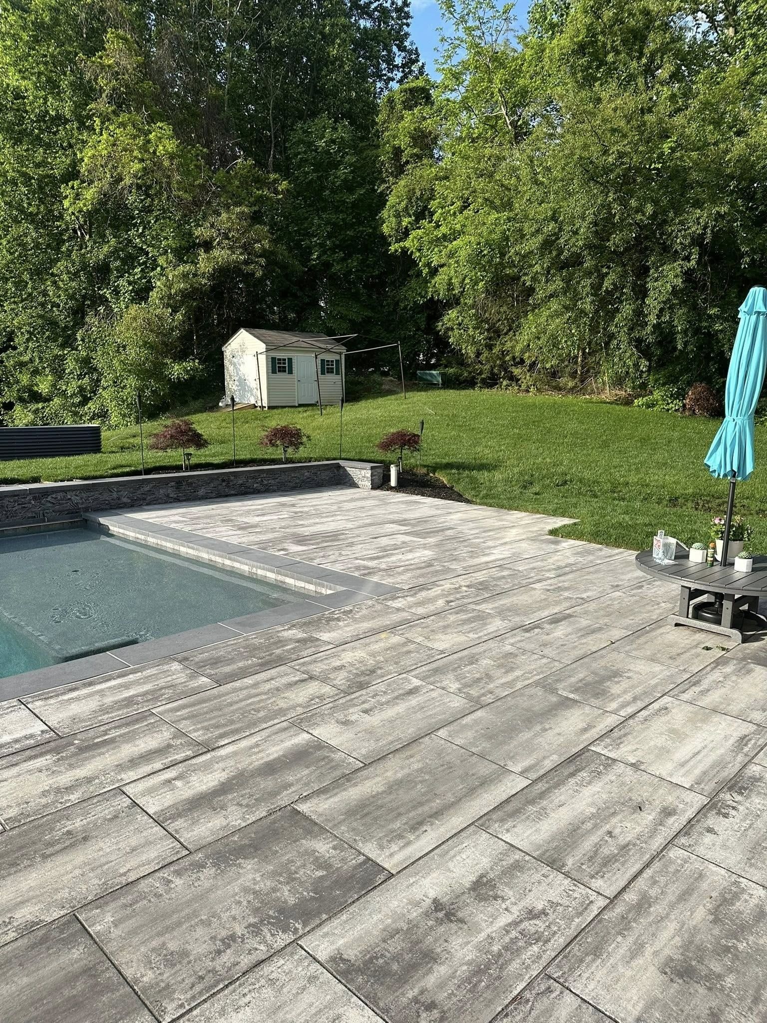 Patio with a pool, umbrella, and small shed on a grassy hill.