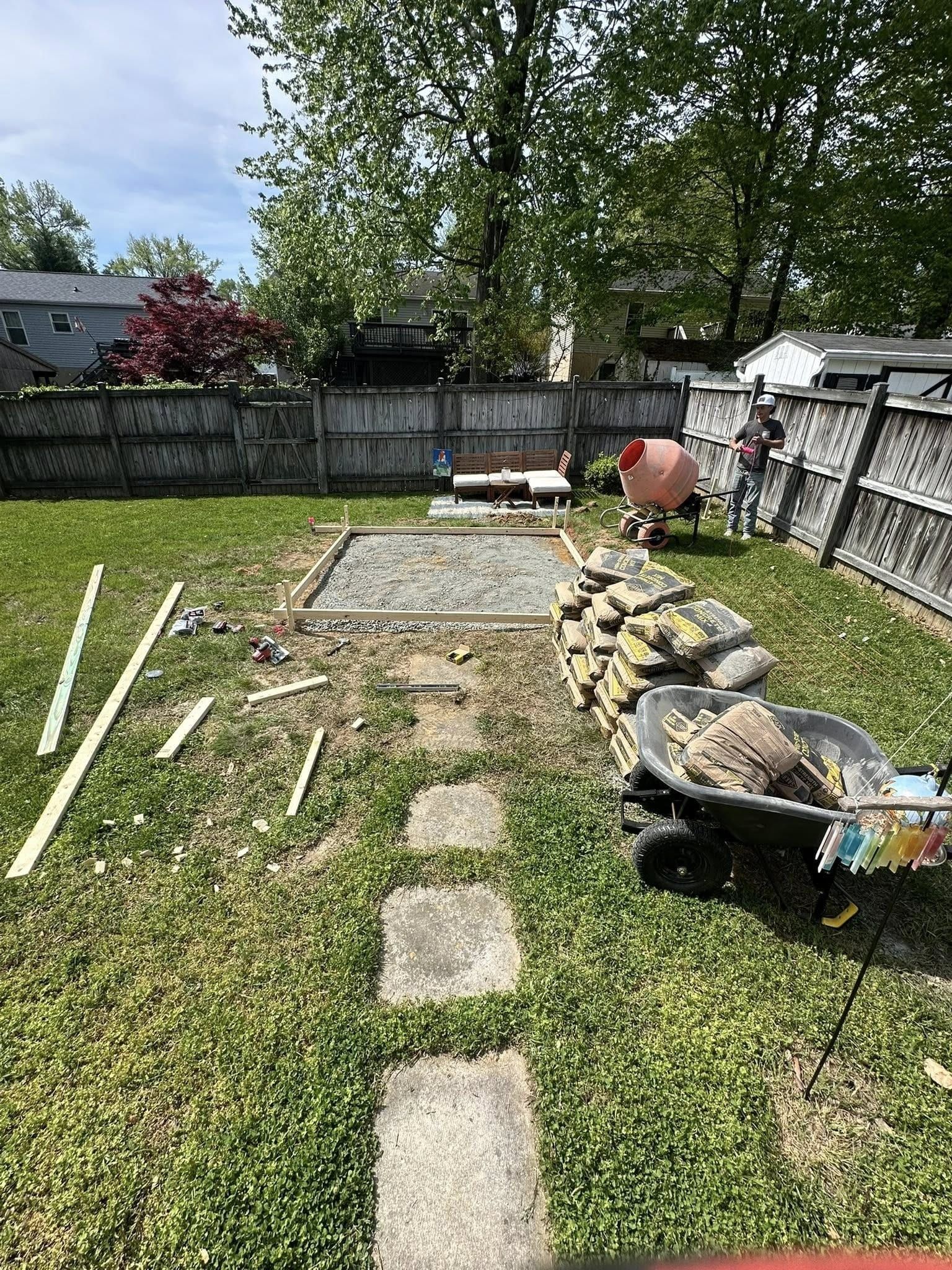 Backyard with gravel base, pavers path, concrete mixer, person, and materials for construction.