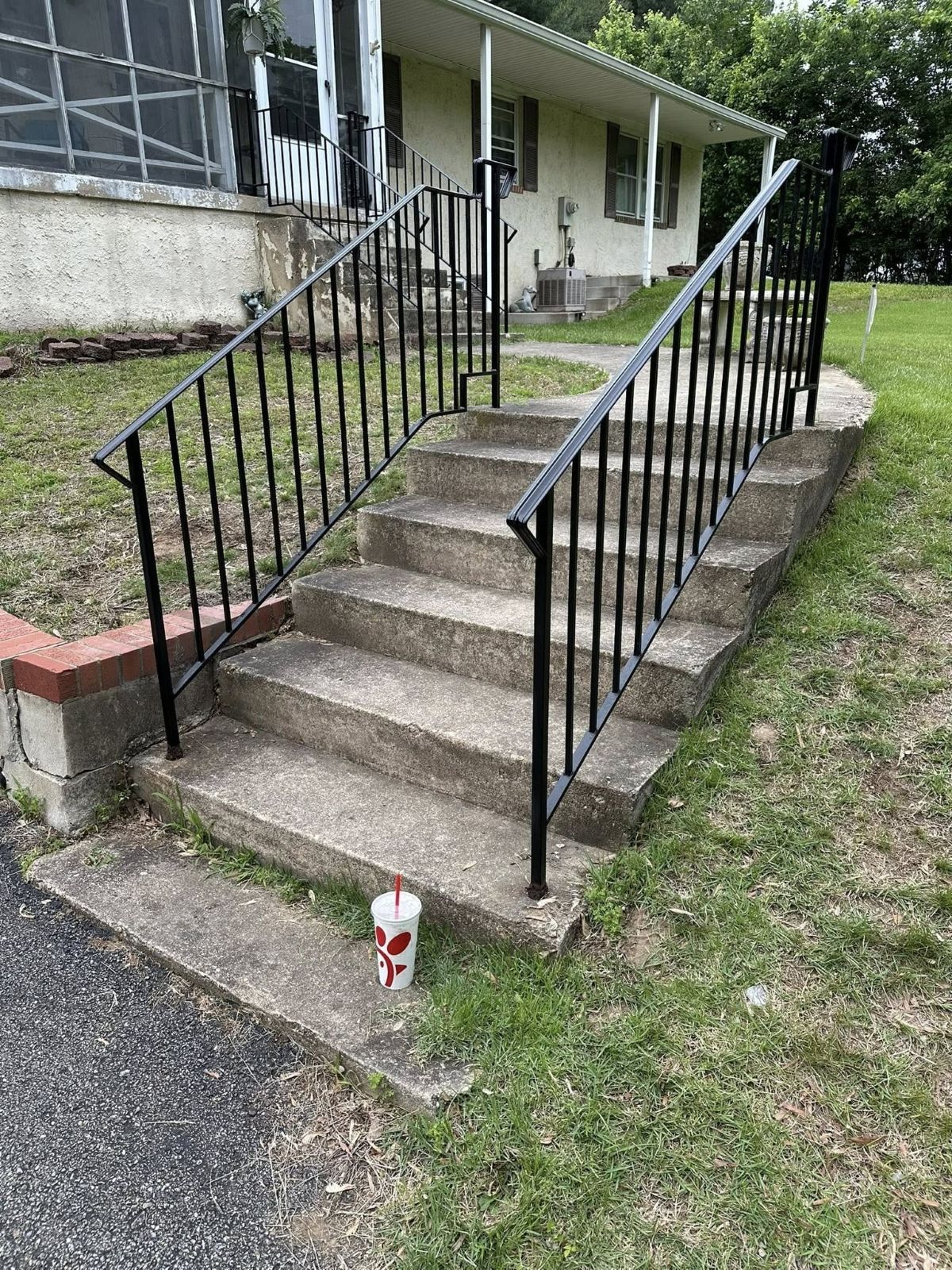 Concrete steps with black metal railings leading to a light-colored house with green grass. A drink cup sits on a step.
