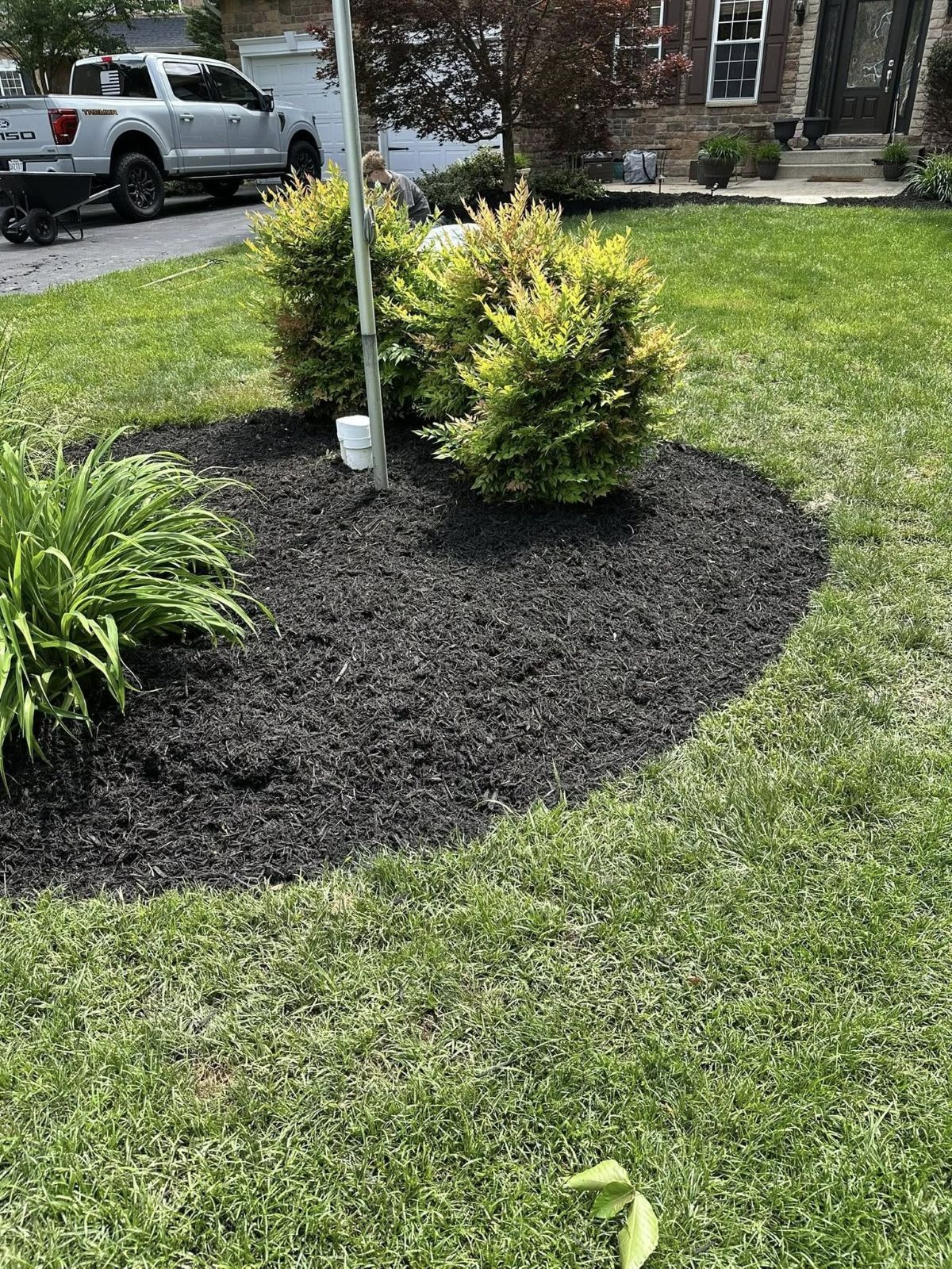 Black mulch surrounds green bushes and a flagpole in a lawn. A truck is parked in the background.
