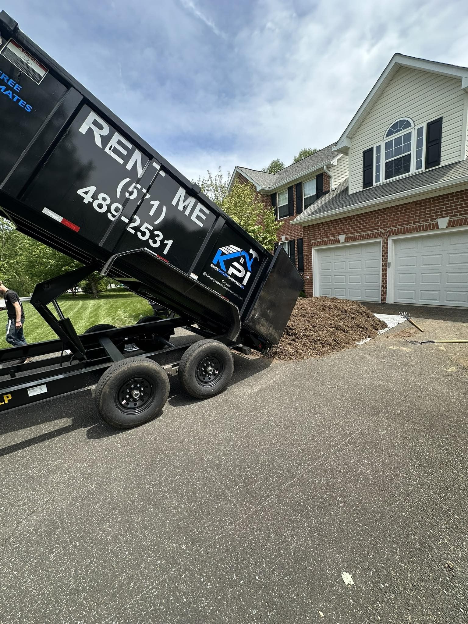 A black rental dump trailer unloading mulch onto a driveway in front of a house.
