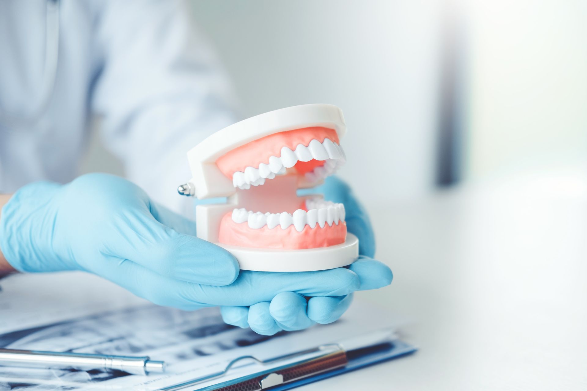 Gloved hands holding a dental model with open pink gums and white teeth on a clipboard in a clinic.