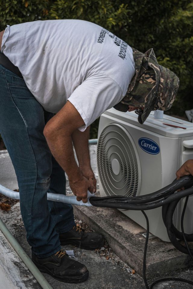 Person in white shirt and hat connecting pipes to an air conditioning unit outdoors.