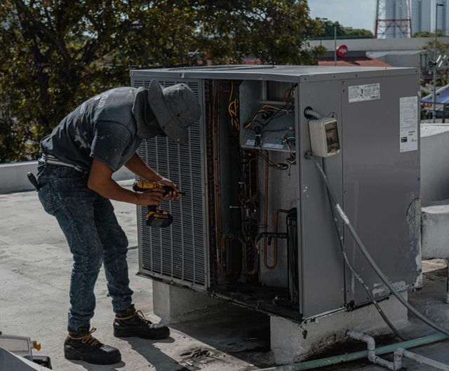 Person repairs an air conditioner on a rooftop, using a drill. Gray unit with open panel, blue jeans, and gray hat.