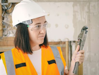 Woman in work gear, safety glasses, and hard hat holding a wrench; looking at it skeptically in a construction setting.