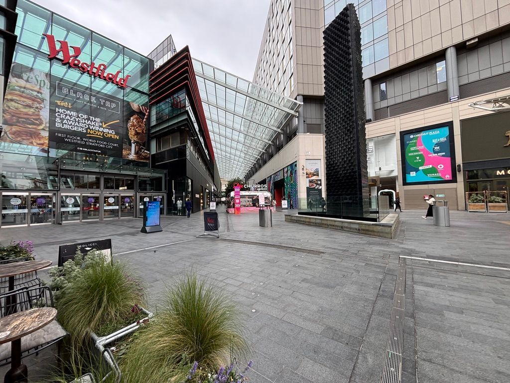 The Westfield shopping center's outdoor plaza, featuring modern architecture, a black sculptural monument, and a storefront.