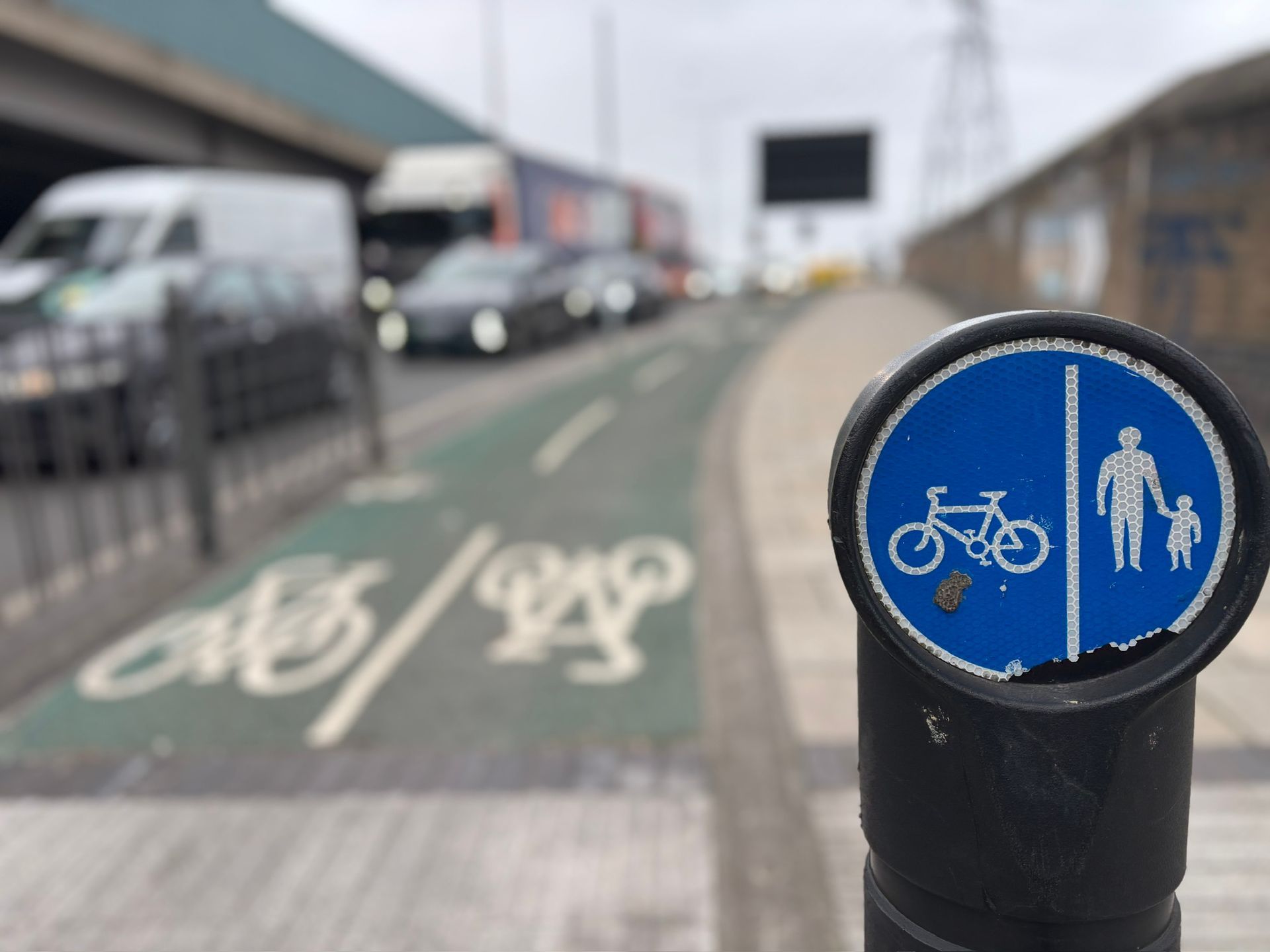 A blue, circular sign shows a bicycle and two pedestrians, marking a shared path beside a road with cars and a truck.