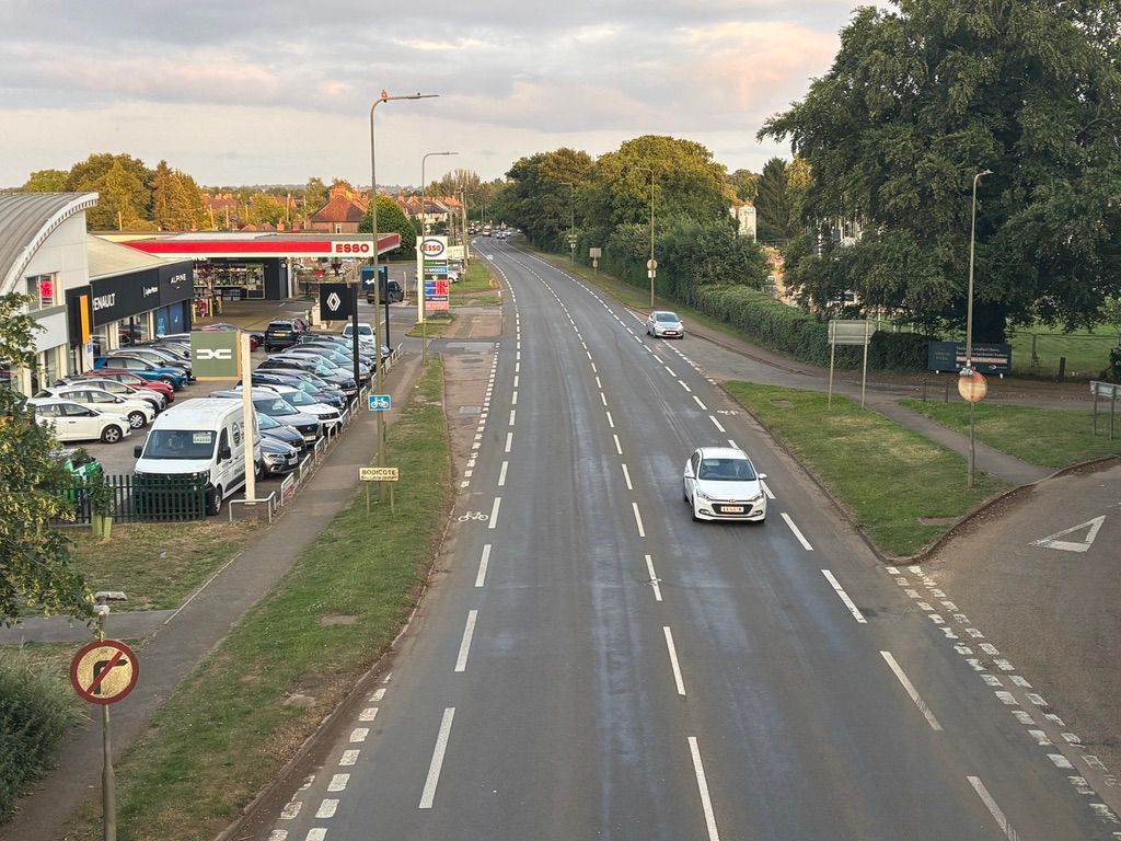 A road view with a gas station and parked cars on the left, a driving white car, and surrounding trees.