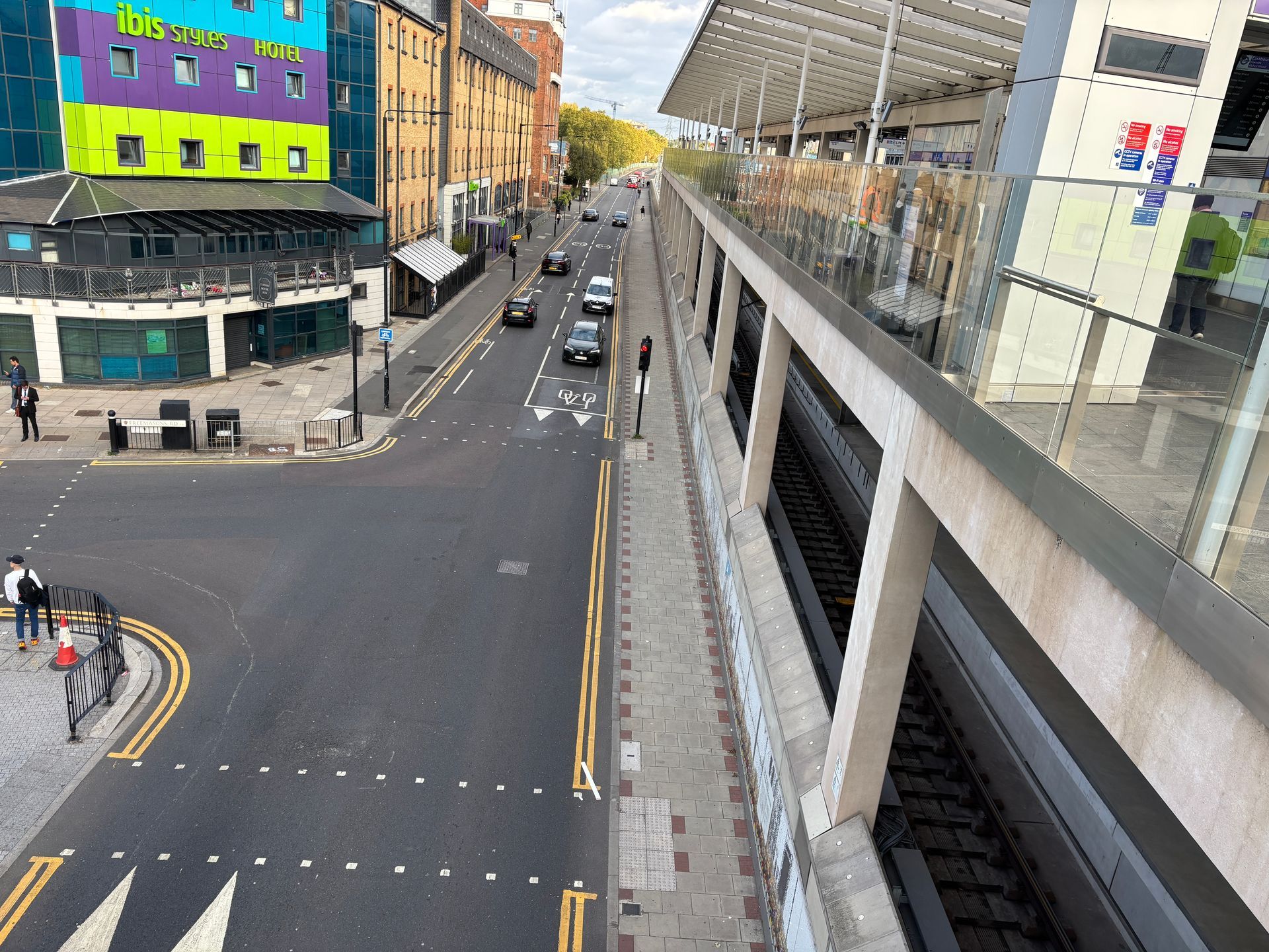A city street runs alongside a raised train track platform with an ibis hotel visible in the background.