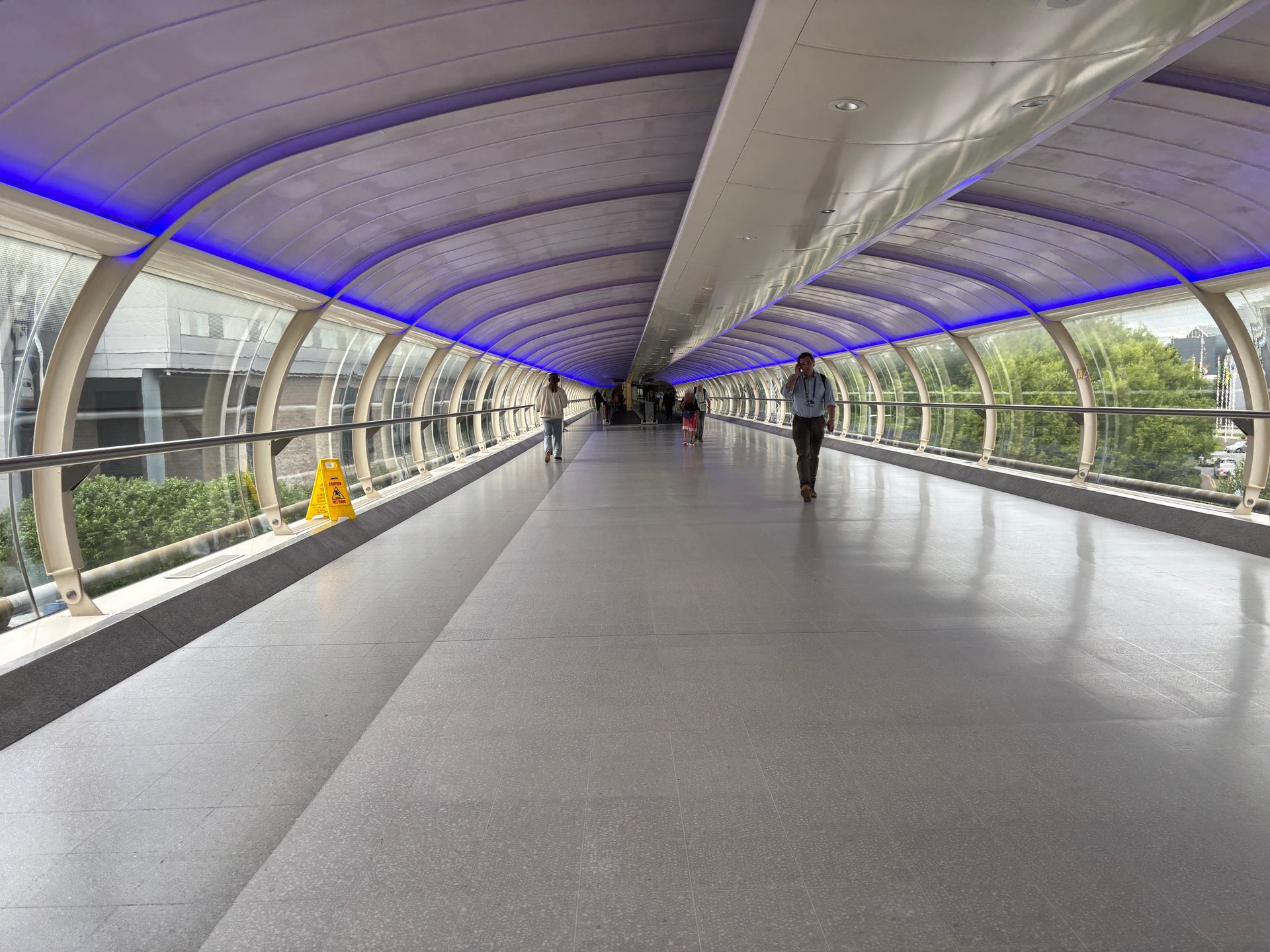 A long, indoor pedestrian walkway with purple ceiling lighting, glass walls, and a few people walking through.
