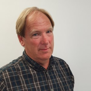 A fair-haired person wearing a plaid button-down shirt stands against a plain white background, looking toward the camera.