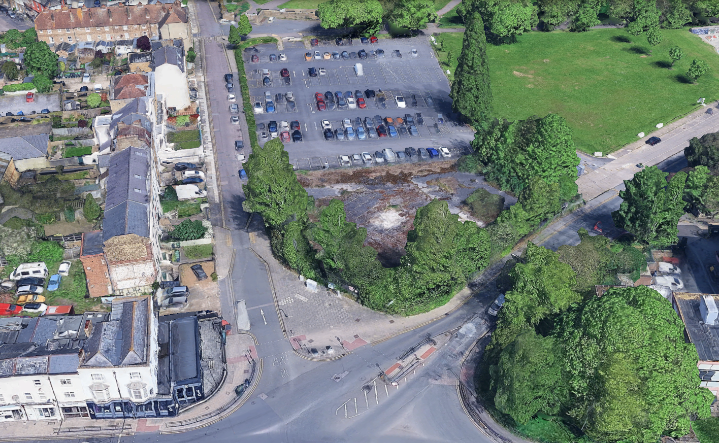 Aerial view of a parking lot bordered by a street, residential buildings, and a patch of green space with trees.