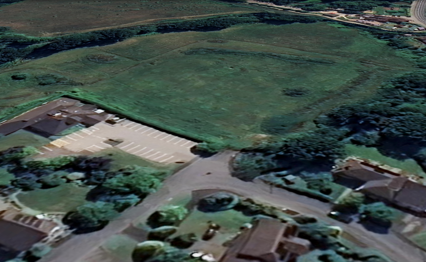 Aerial view of a parking lot bordered by a large, open green field and surrounding residential houses with trees.
