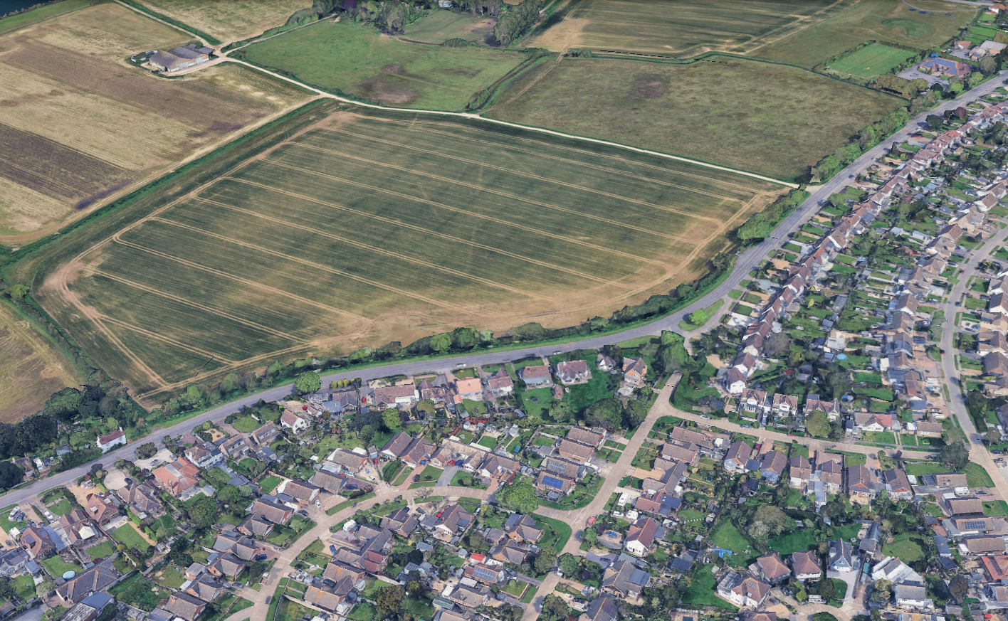 An aerial view of a residential neighborhood bordering a large, rectangular agricultural field with visible crop rows.