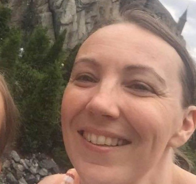 A close-up selfie of a smiling person outdoors, with a rocky, castle-like structure in the background.