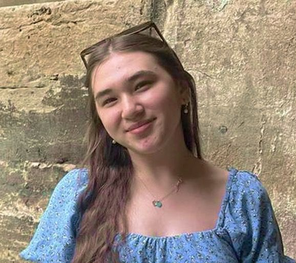 A person with long brown hair wearing a blue floral top and a necklace, smiling against a textured stone wall.