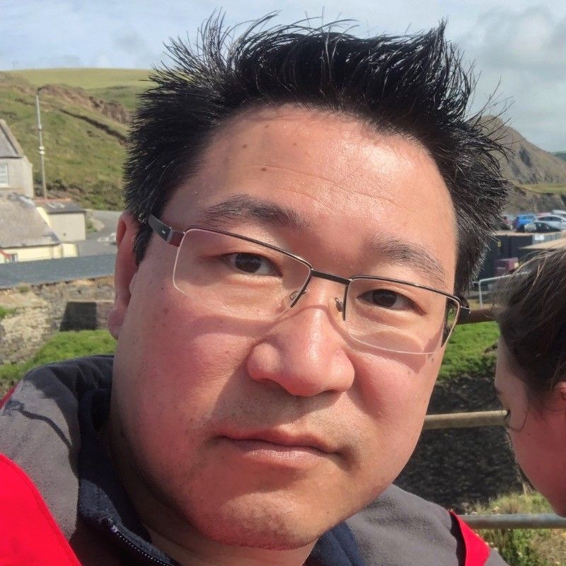 A man with short dark hair and glasses poses for a selfie outdoors, with rolling hills and a building in the background.