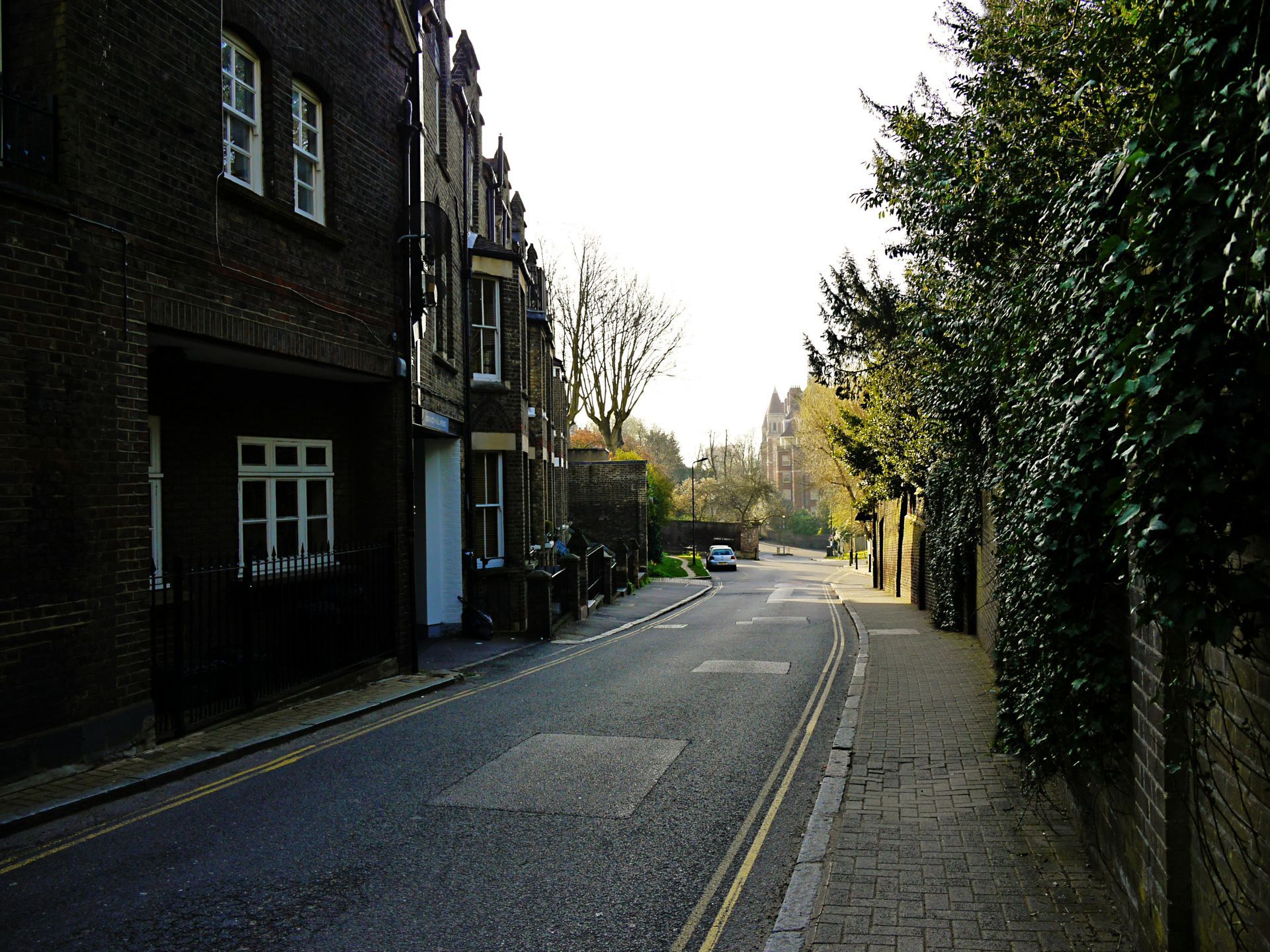 A narrow, paved street lined with dark brick buildings on the left and a stone wall with greenery on the right.