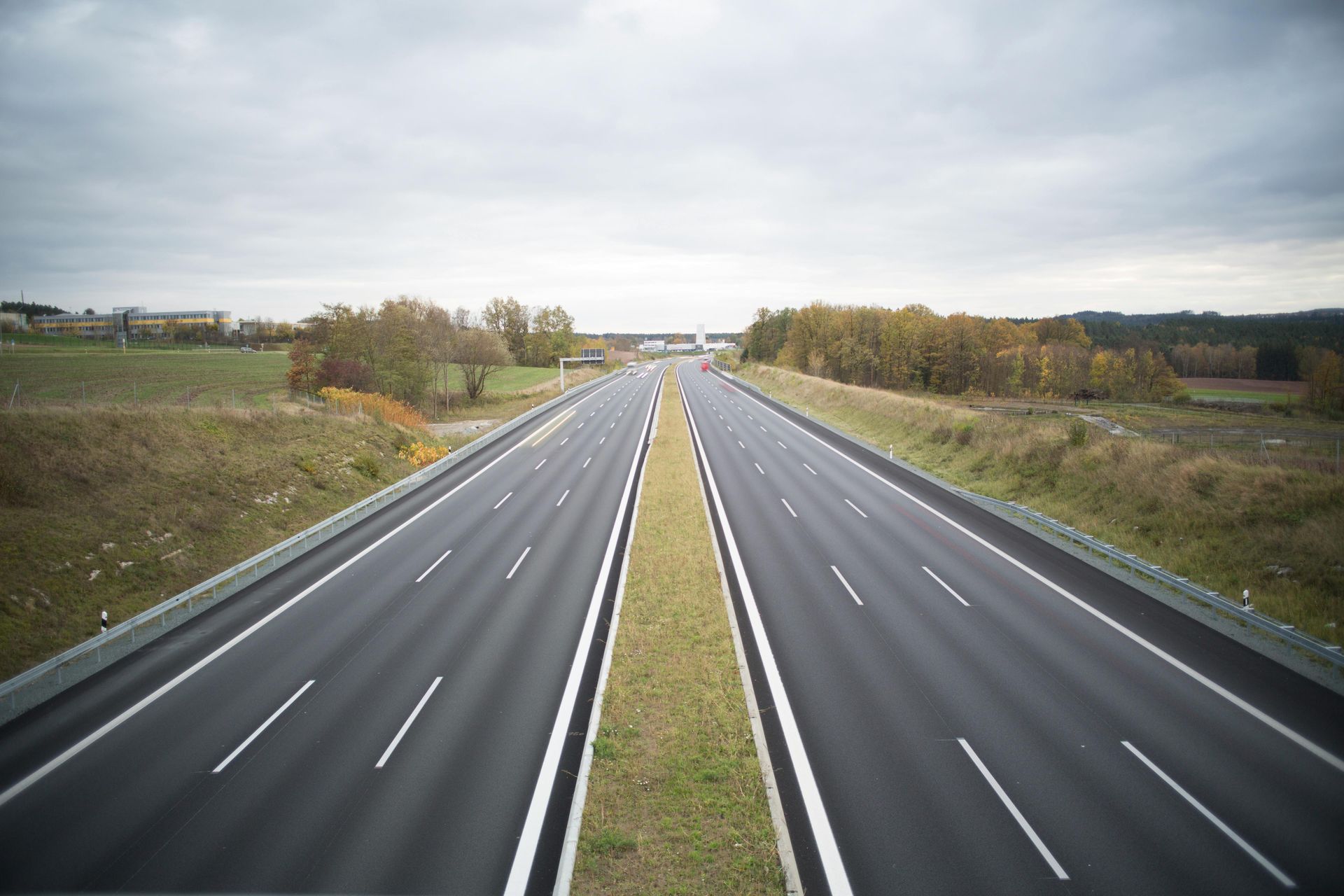A wide, empty dual-carriageway highway stretching into the distance through a rural, autumn landscape under a cloudy sky.