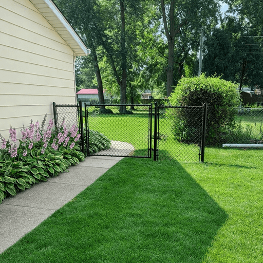 A black chain link fence encloses a green lawn, with a concrete path leading to a gate and the side of a house.