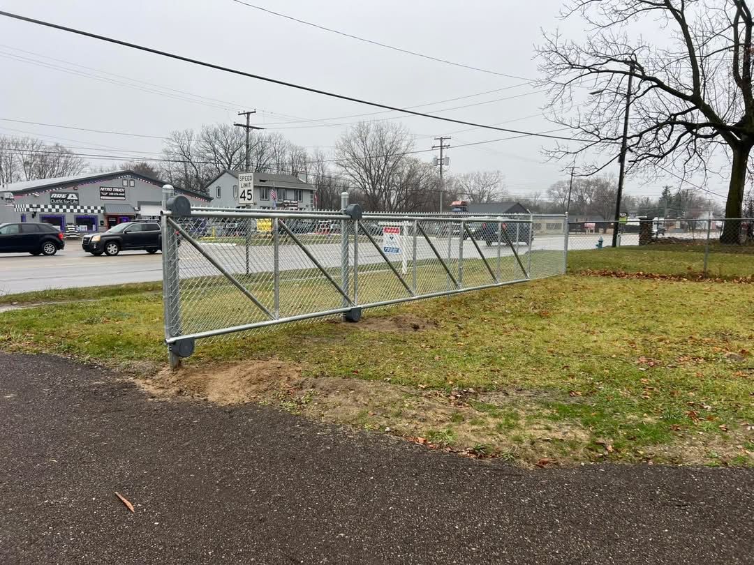 Chain-link fence borders a grassy area next to a paved path, with cars and buildings in the background on an overcast day.