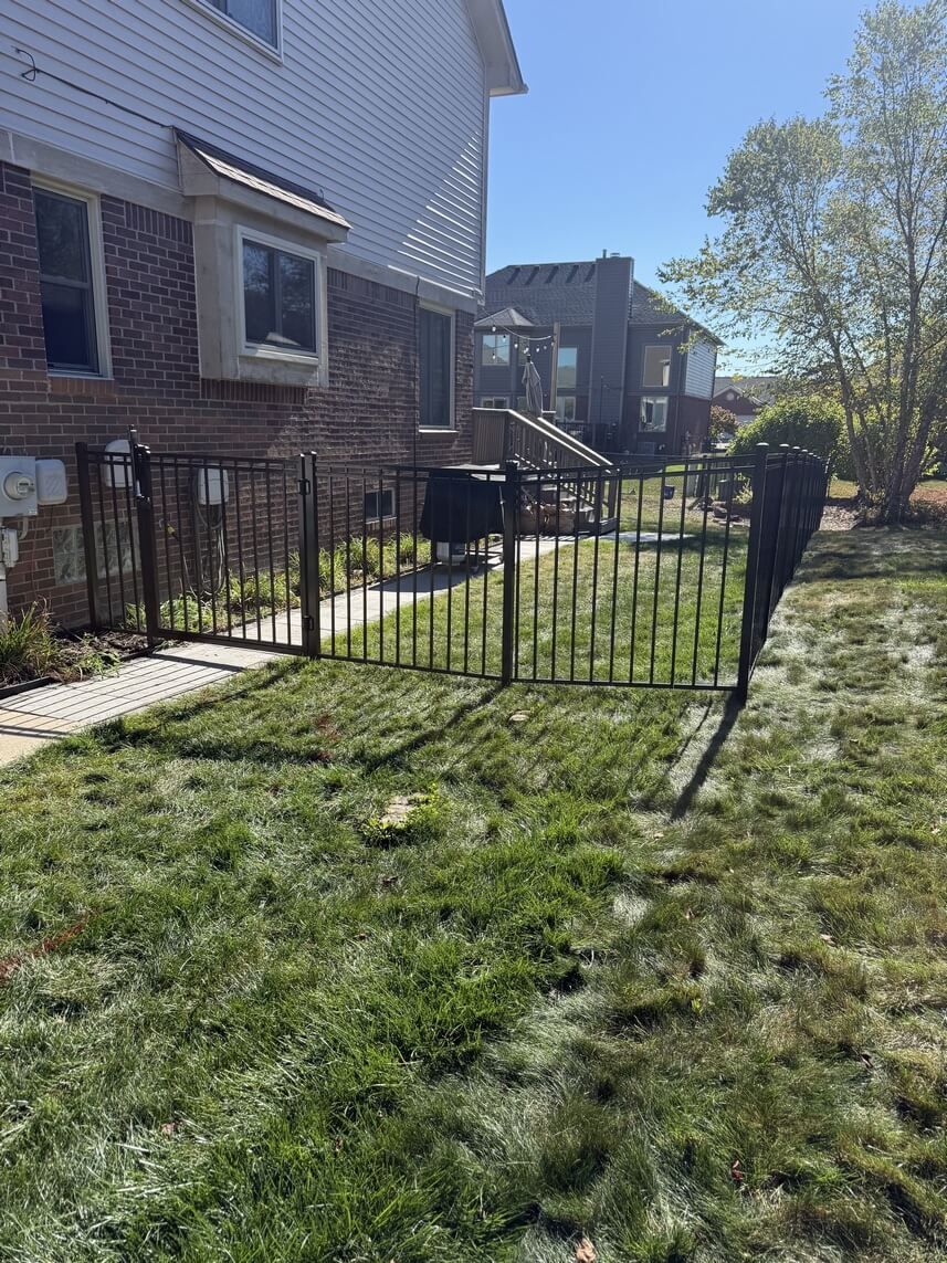 A black metal fence surrounds a grassy backyard, adjacent to a brick house on a sunny day.