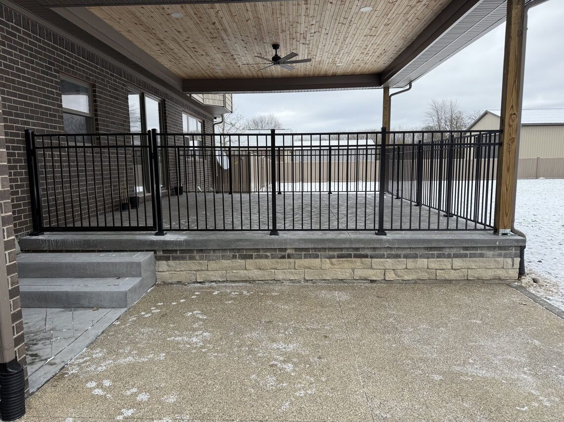 Covered patio with black railing, light-colored brick facade, stone steps, and gravel ground.