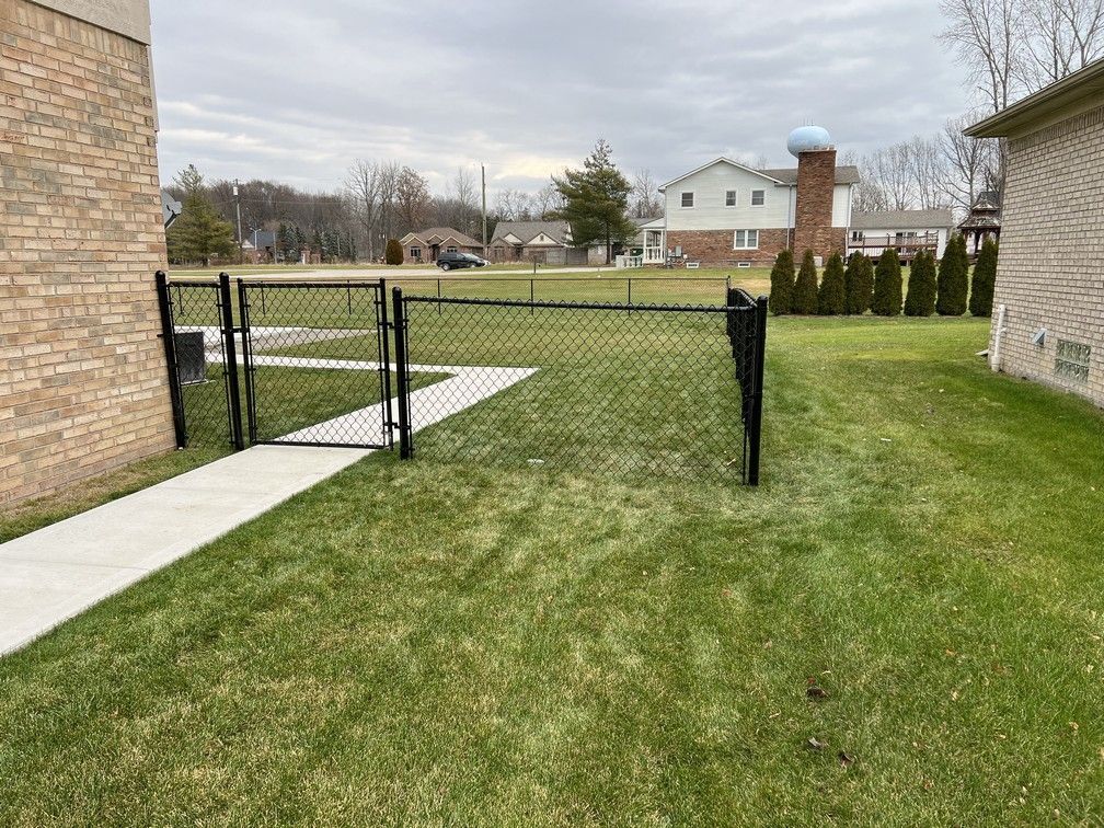 A chain link fence surrounds a lush green yard next to a brick building.
