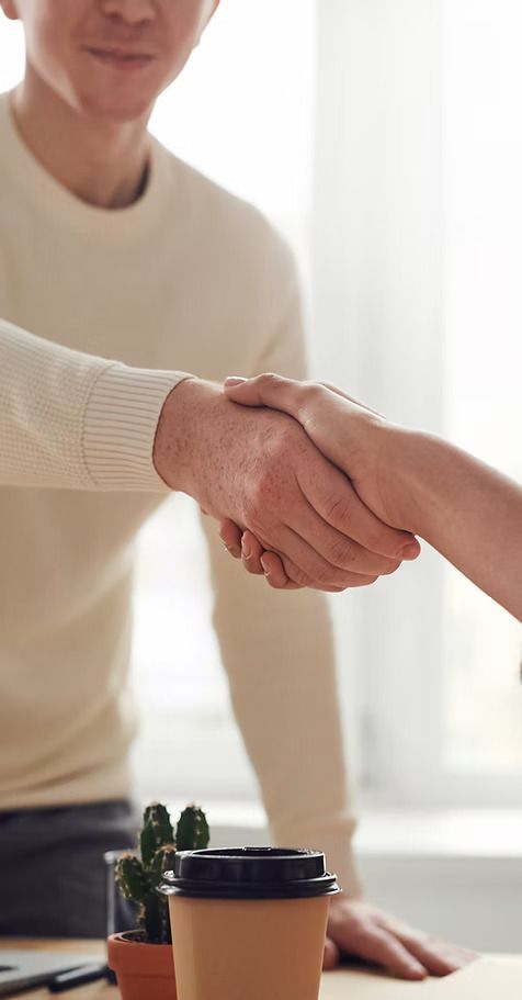 Two people shaking hands over a desk, one with a neutral expression in a bright room.