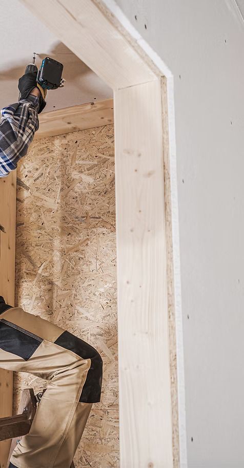 Person using a power drill to install wooden door frame. Inside a construction setting.
