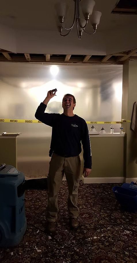 Man in a room with a damaged ceiling holds an object up, smiling. The room has plastic sheeting and a blue device.