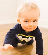 Baby with blonde hair crawling on a wooden floor, wearing a dark blue shirt with a yellow design, smiling.