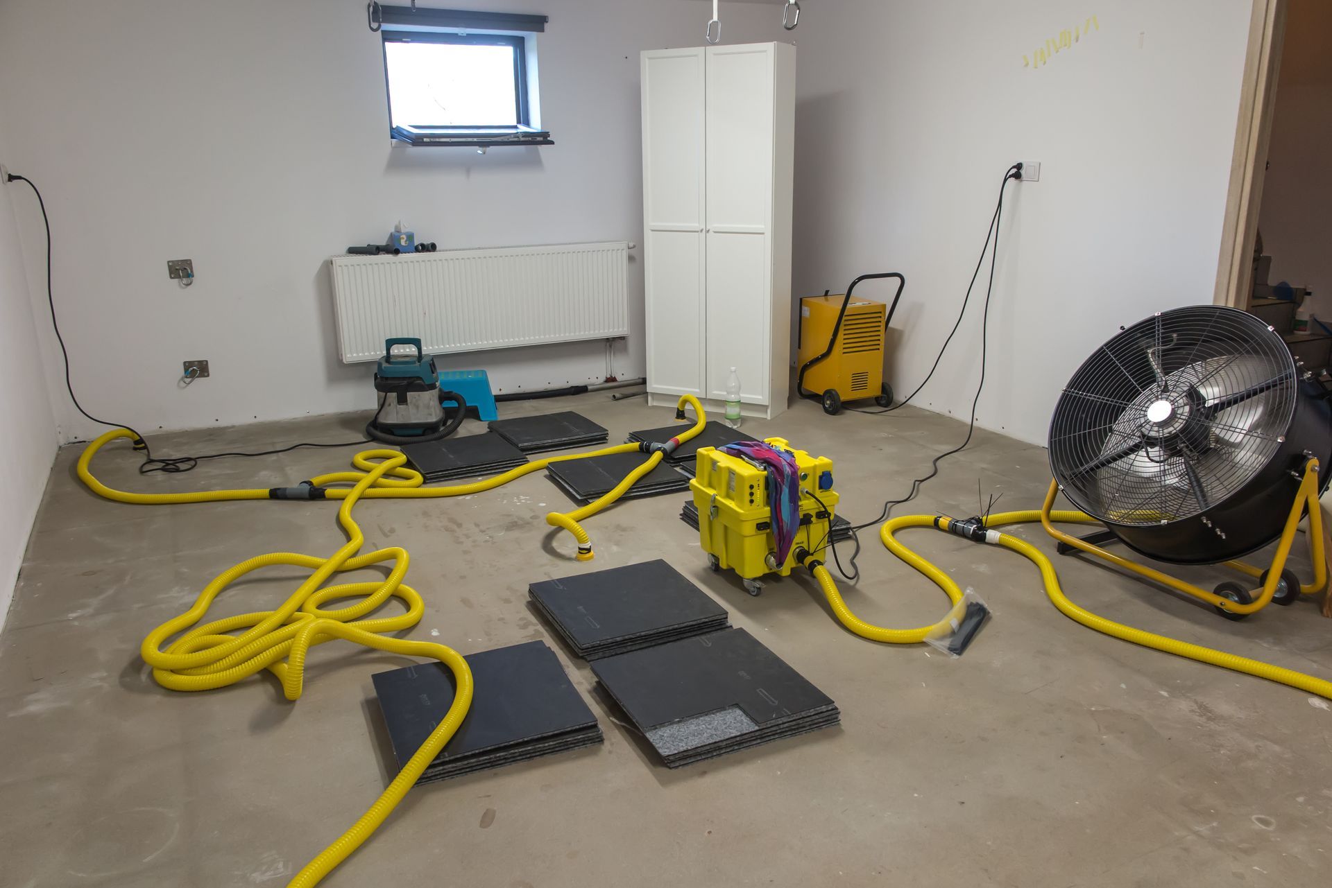 Water damage restoration in a basement room with air movers, dehumidifier, and yellow hoses drying concrete flooring after flooding. Water damage restoration in a basement room with air movers, dehumidifier, and yellow hoses drying concrete flooring after flooding.