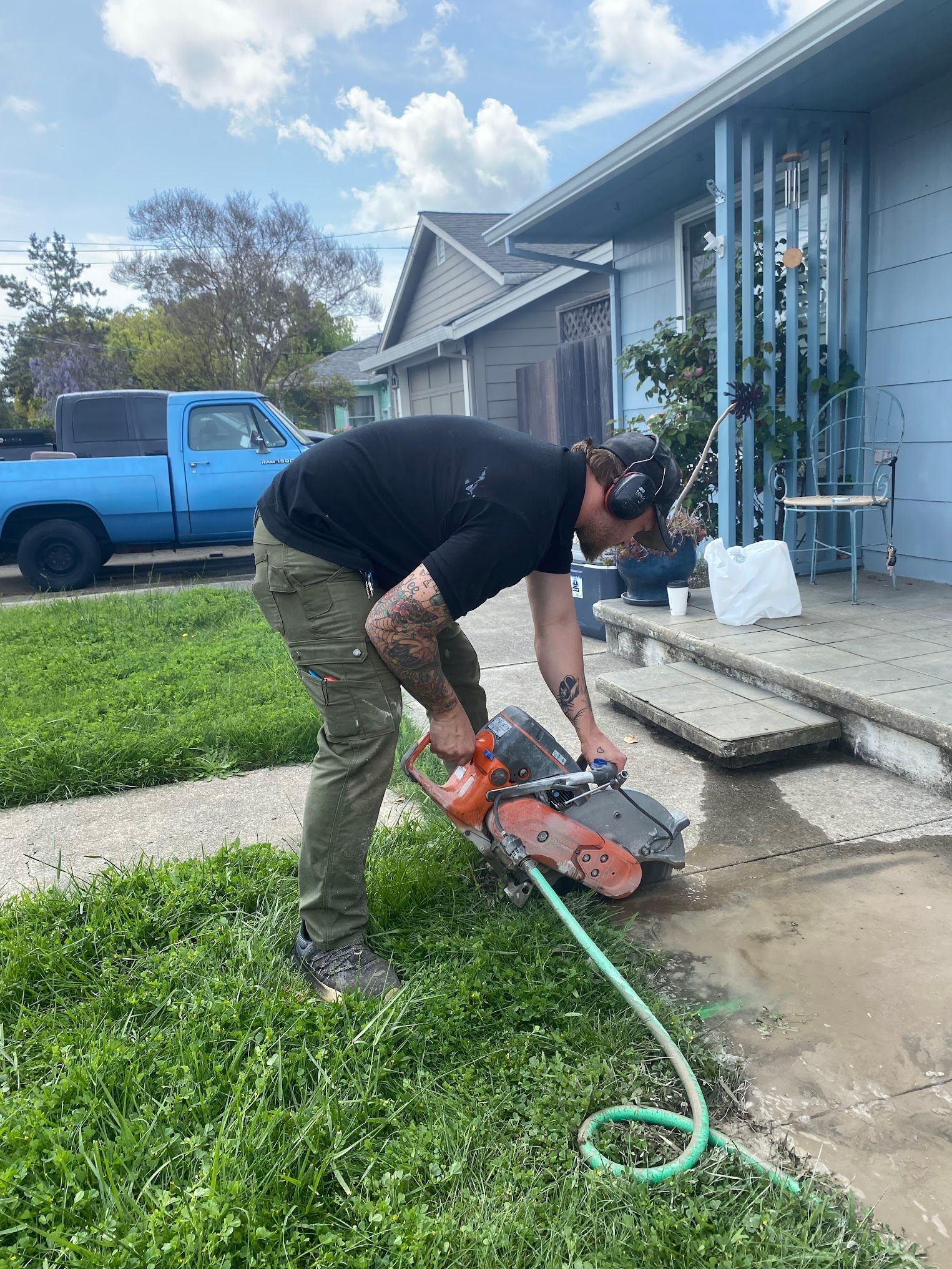 Man cutting concrete with a saw in front of a blue house, green grass, and a blue pickup truck.