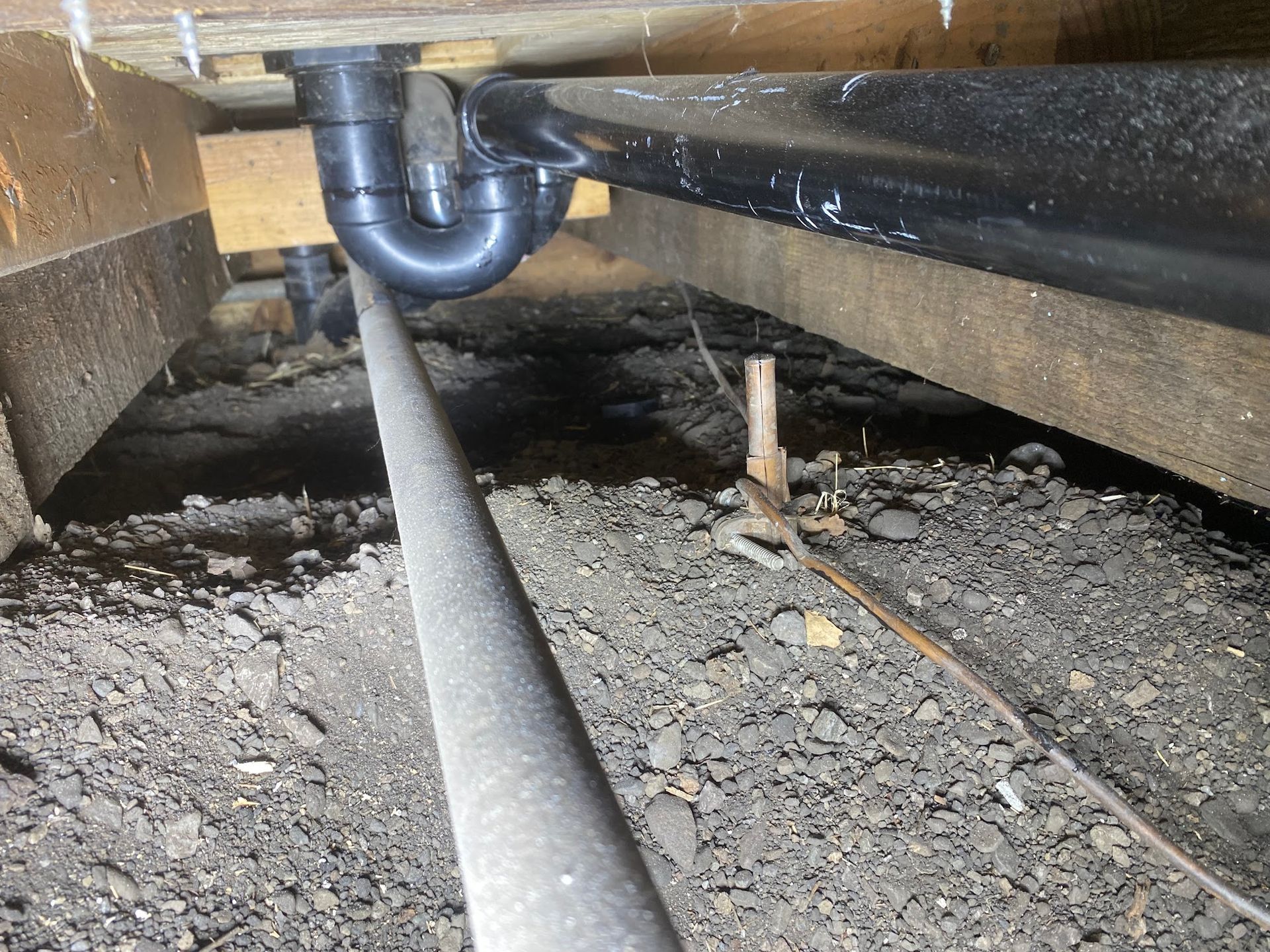 Underside view of plumbing pipes, including a P-trap, under a wooden floor in a crawl space.