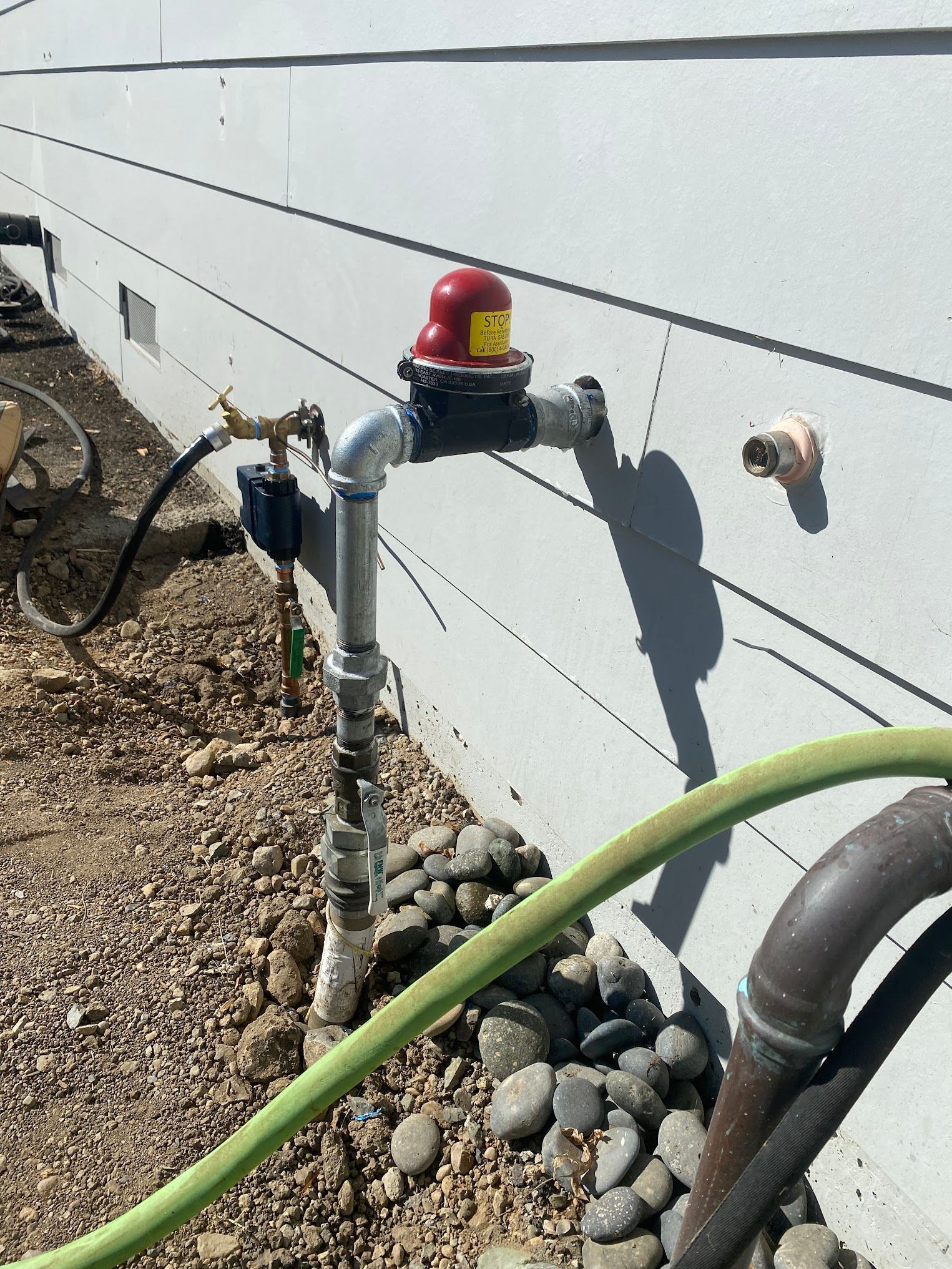 A silver water pipe with a red pressure relief valve attached to a white house. A green hose is connected to the pipe.
