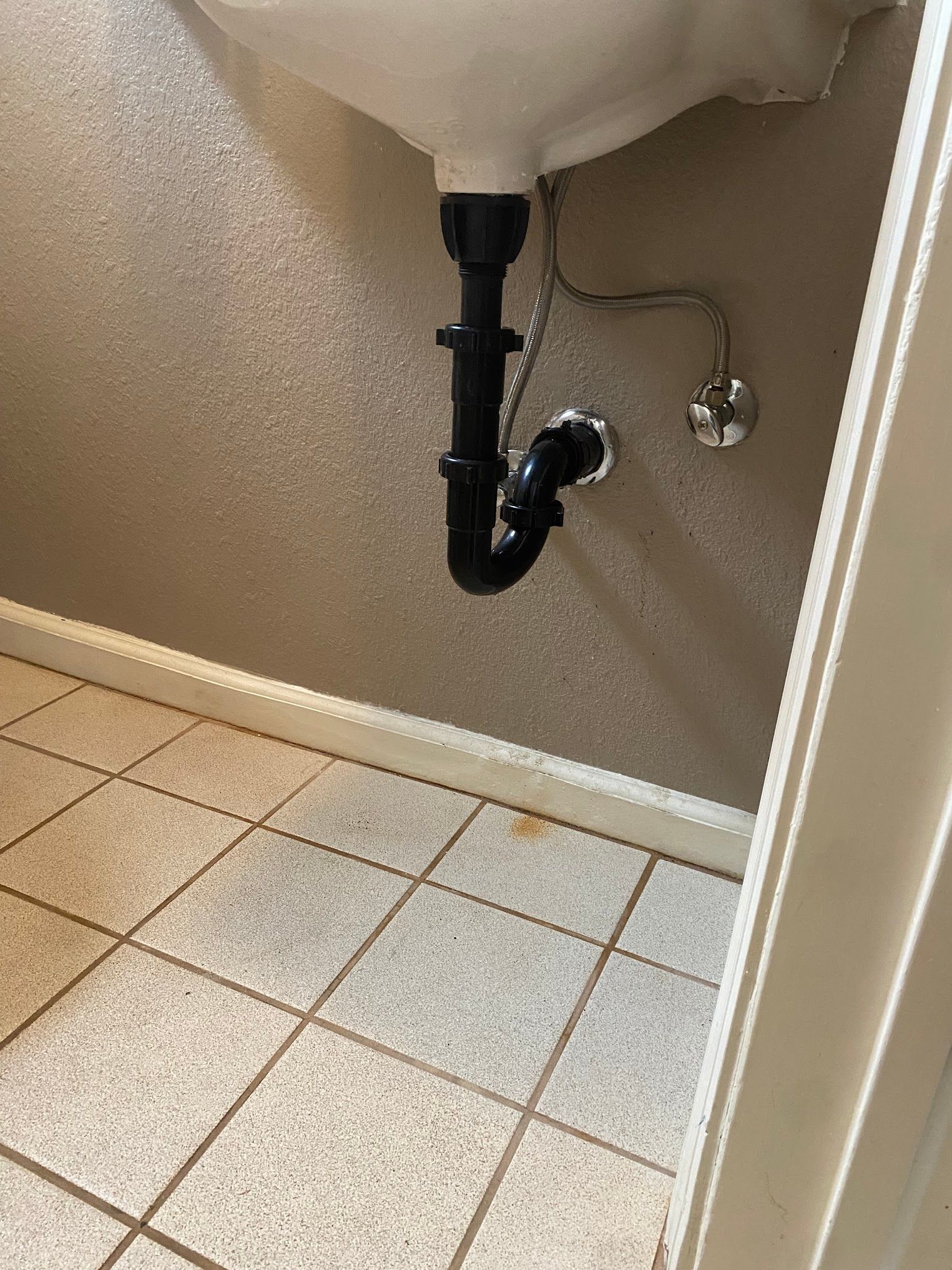 Underside of a white sink with black plumbing against a light brown wall. Tiled floor with baseboard trim.