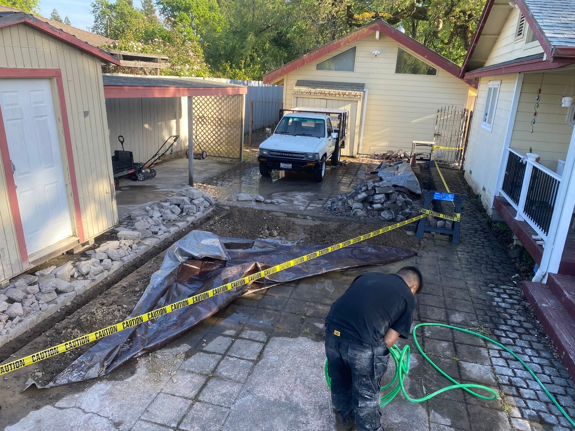 Man working on patio renovation; truck, tools, buildings in a backyard setting.