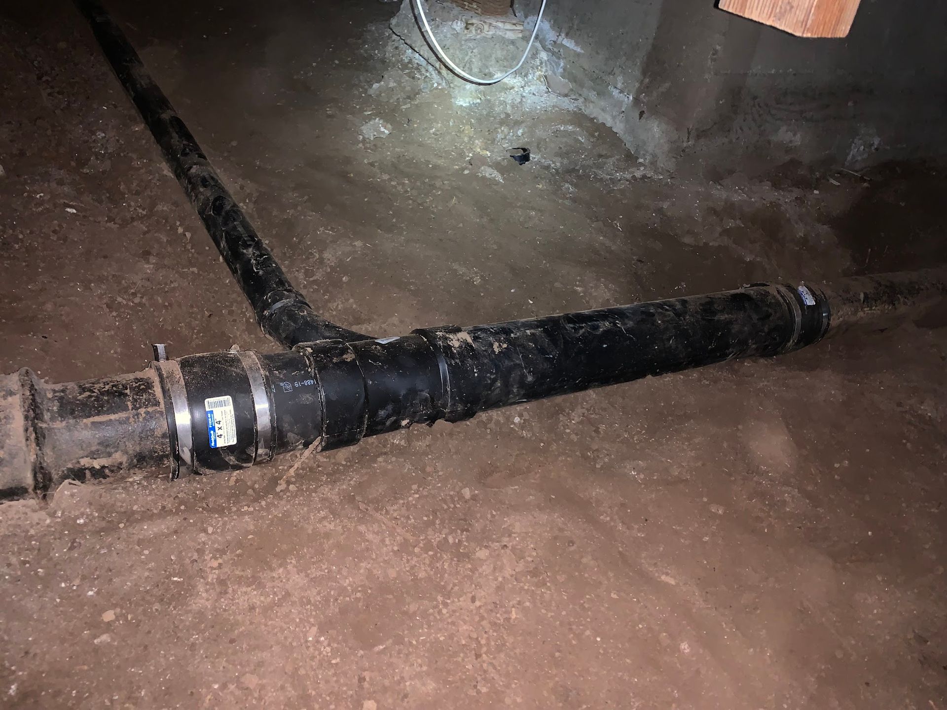 Black plumbing pipes in a crawl space with dirt and debris.