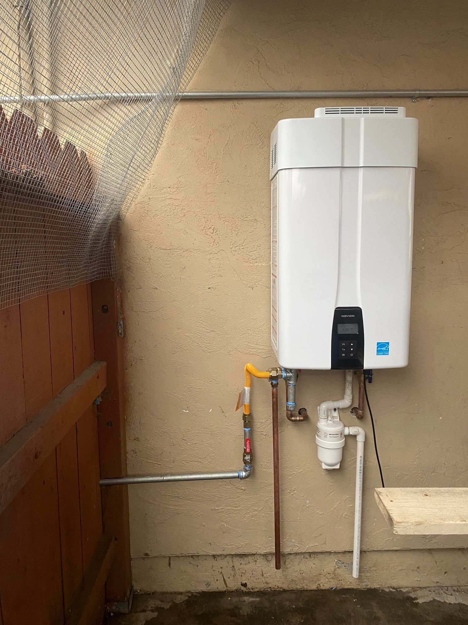 White tankless water heater mounted on a stucco wall, connected to pipes and gas lines.