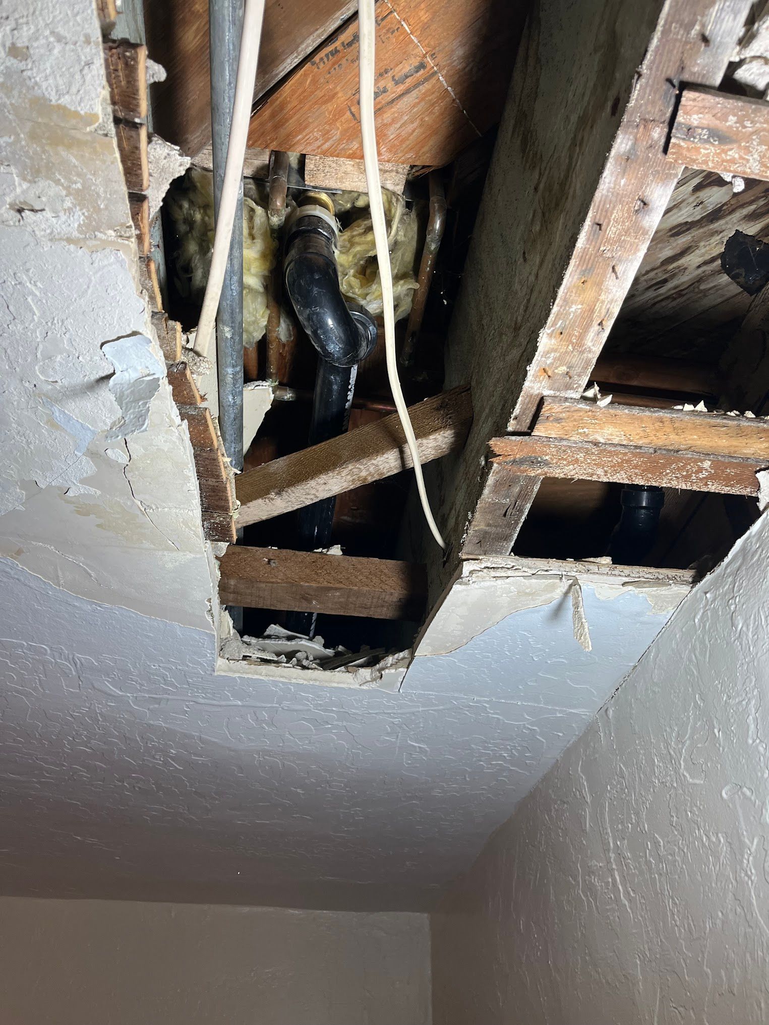 Hole in ceiling reveals plumbing pipes and wooden joists. Insulation and wires are visible.