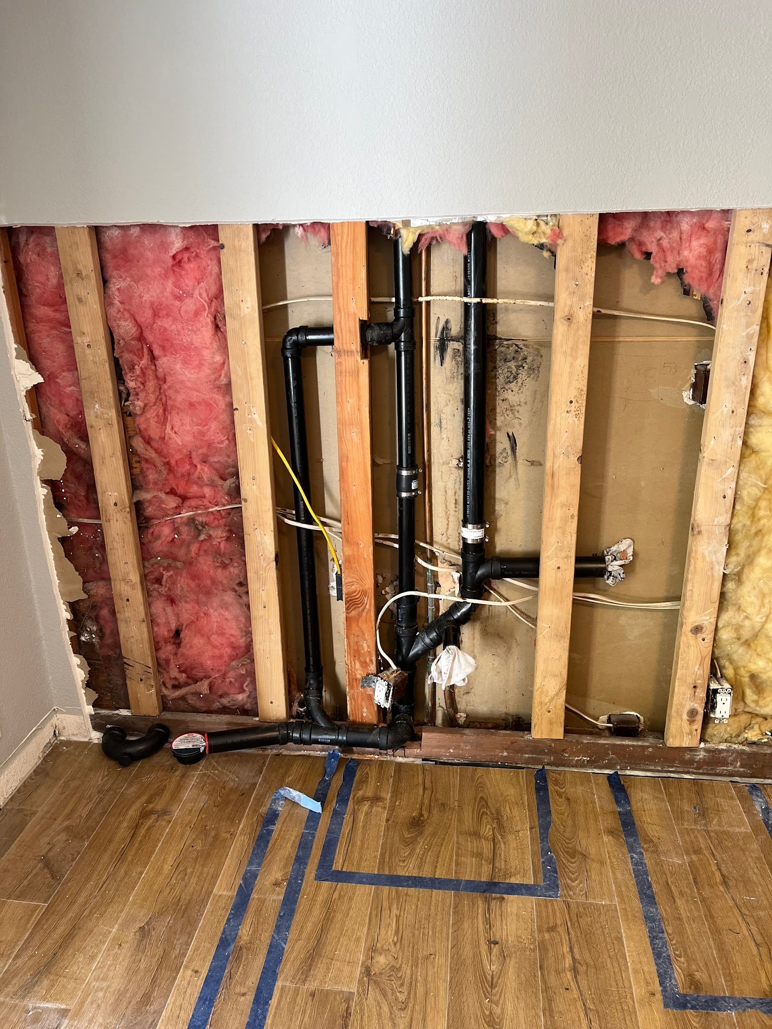 Exposed plumbing inside a wall with black pipes, insulation, and wooden studs, on a wood floor.