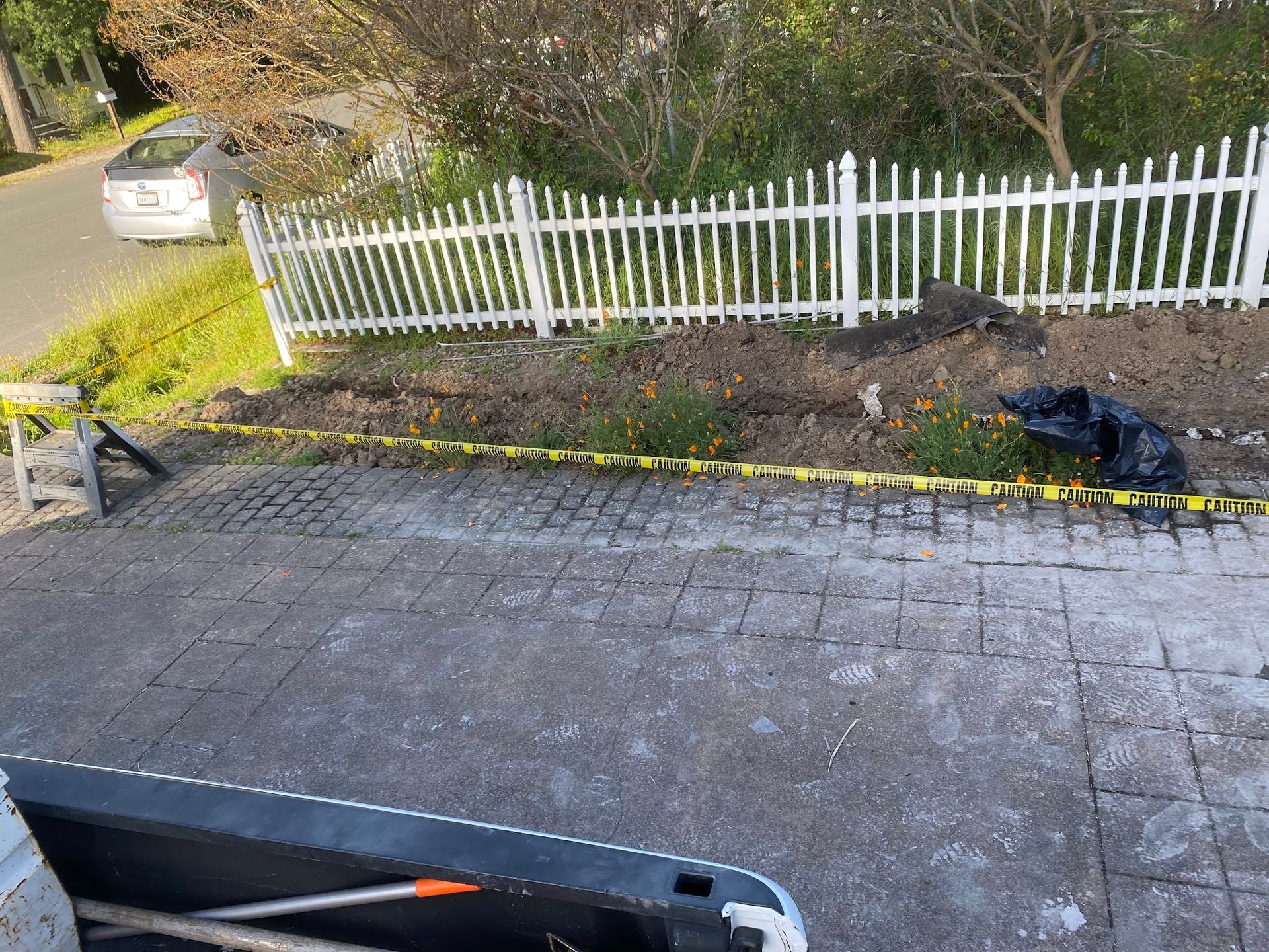 Pavement leading to a white picket fence next to a car on a road. Soil and some flowers are in front of the fence.