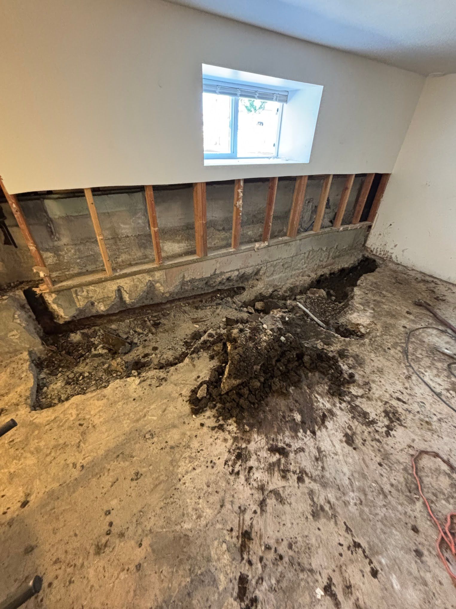 Basement wall under construction, debris scattered. Exposed wood framing, small window, concrete floor.