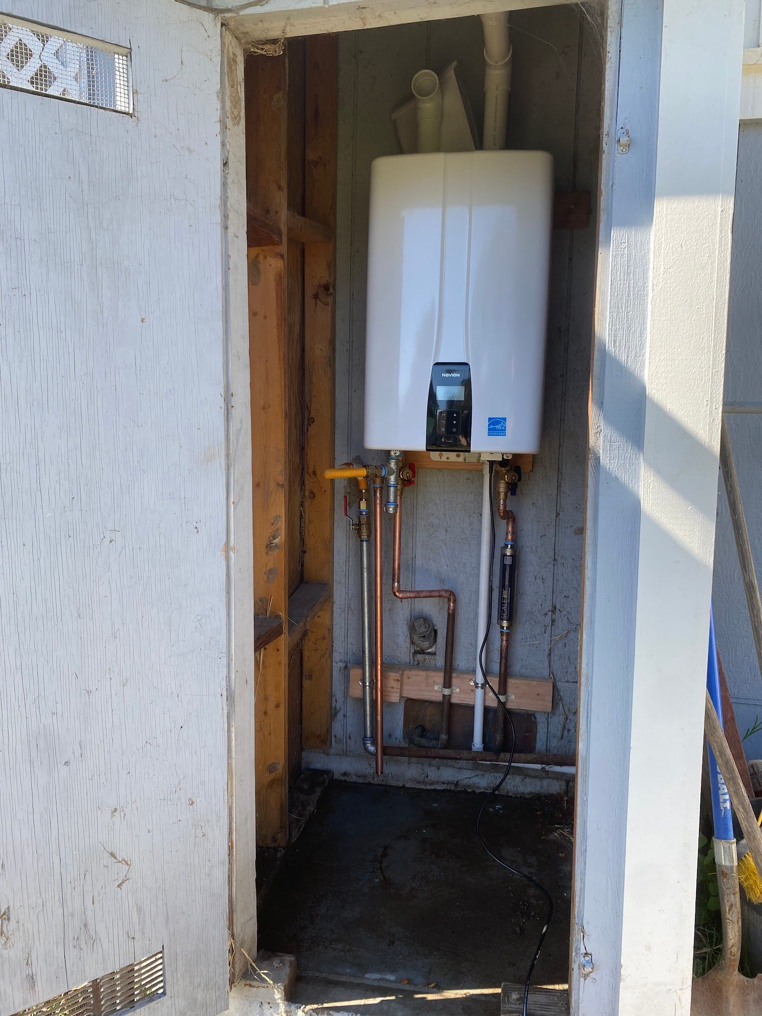 White tank water heater in a narrow outdoor enclosure with plumbing.