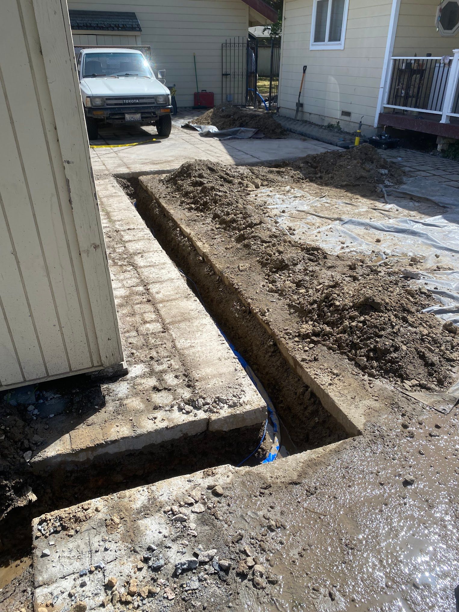 A trench dug in a paved driveway. Dirt piles, a white truck, and buildings are in the background.