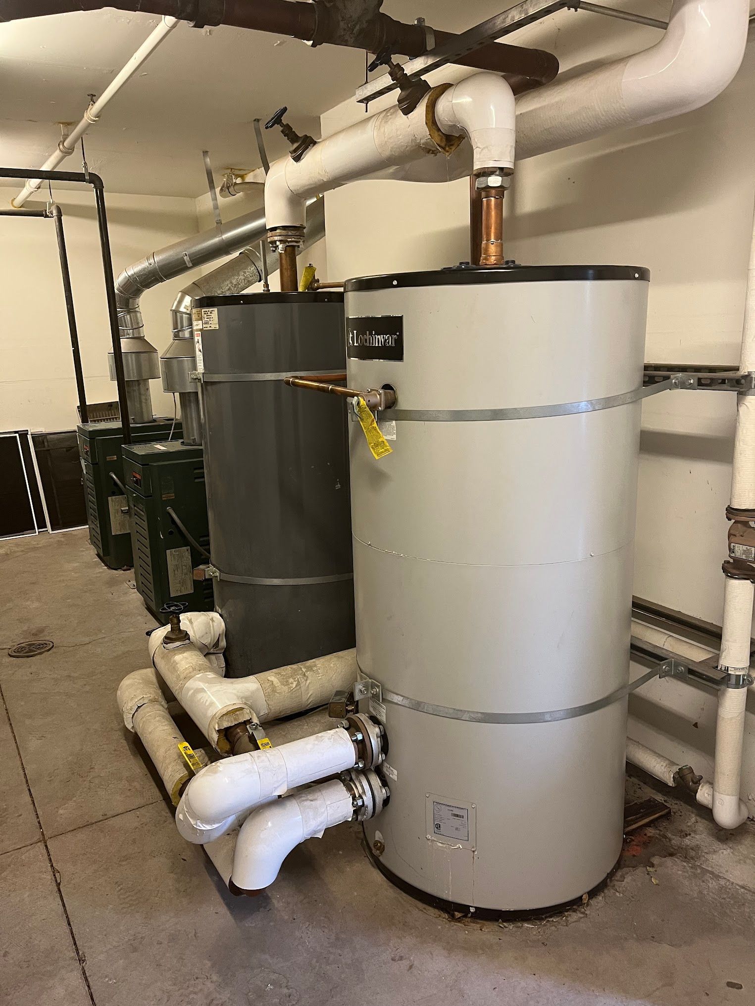 Water heaters in a utility room. Large cylindrical tanks, white and gray, connected by pipes. Concrete floor.